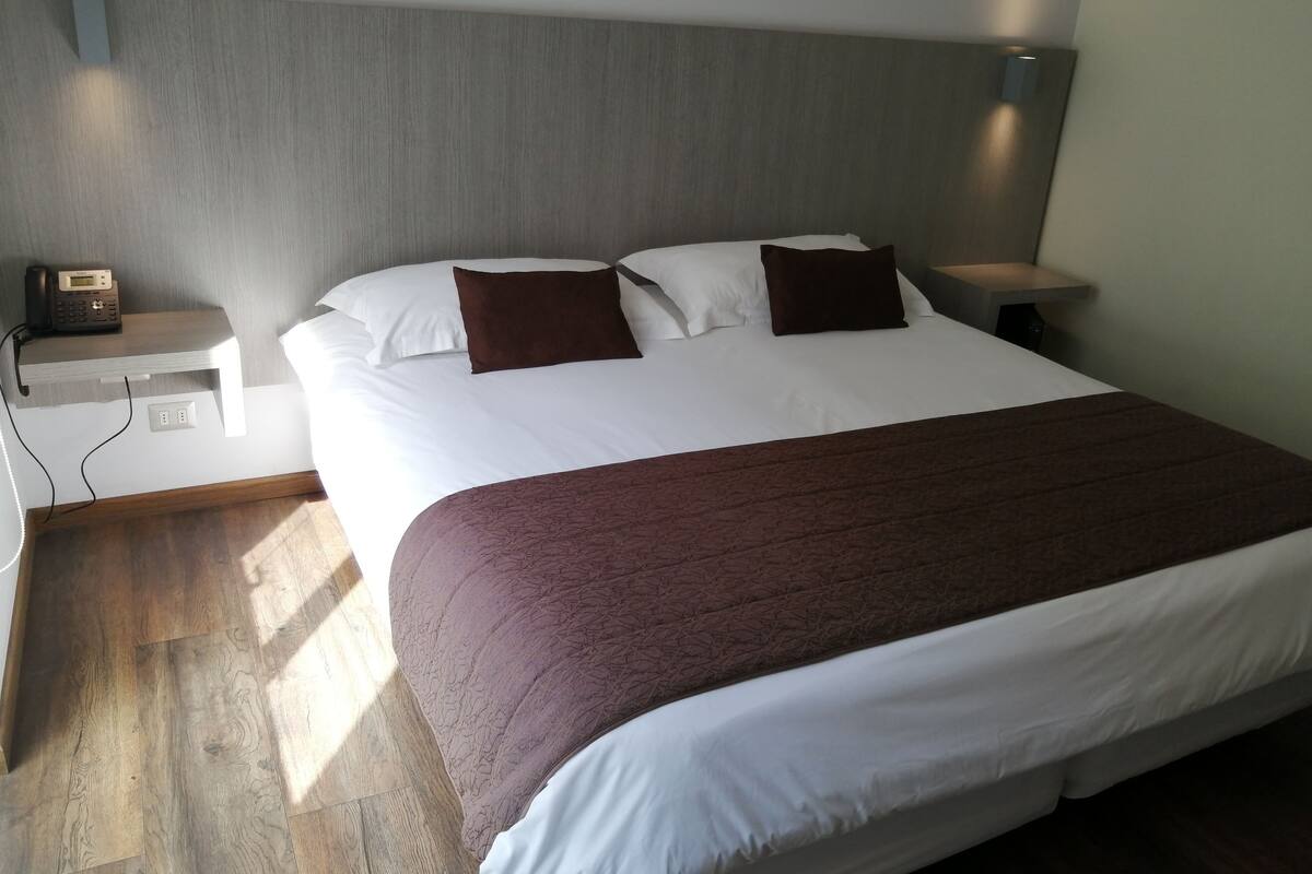 Ein ordentlich bezogenes Bett mit weißer und brauner Tagesdecke, zwei braune Kissen und ein Nachttisch aus Holz mit Telefon.