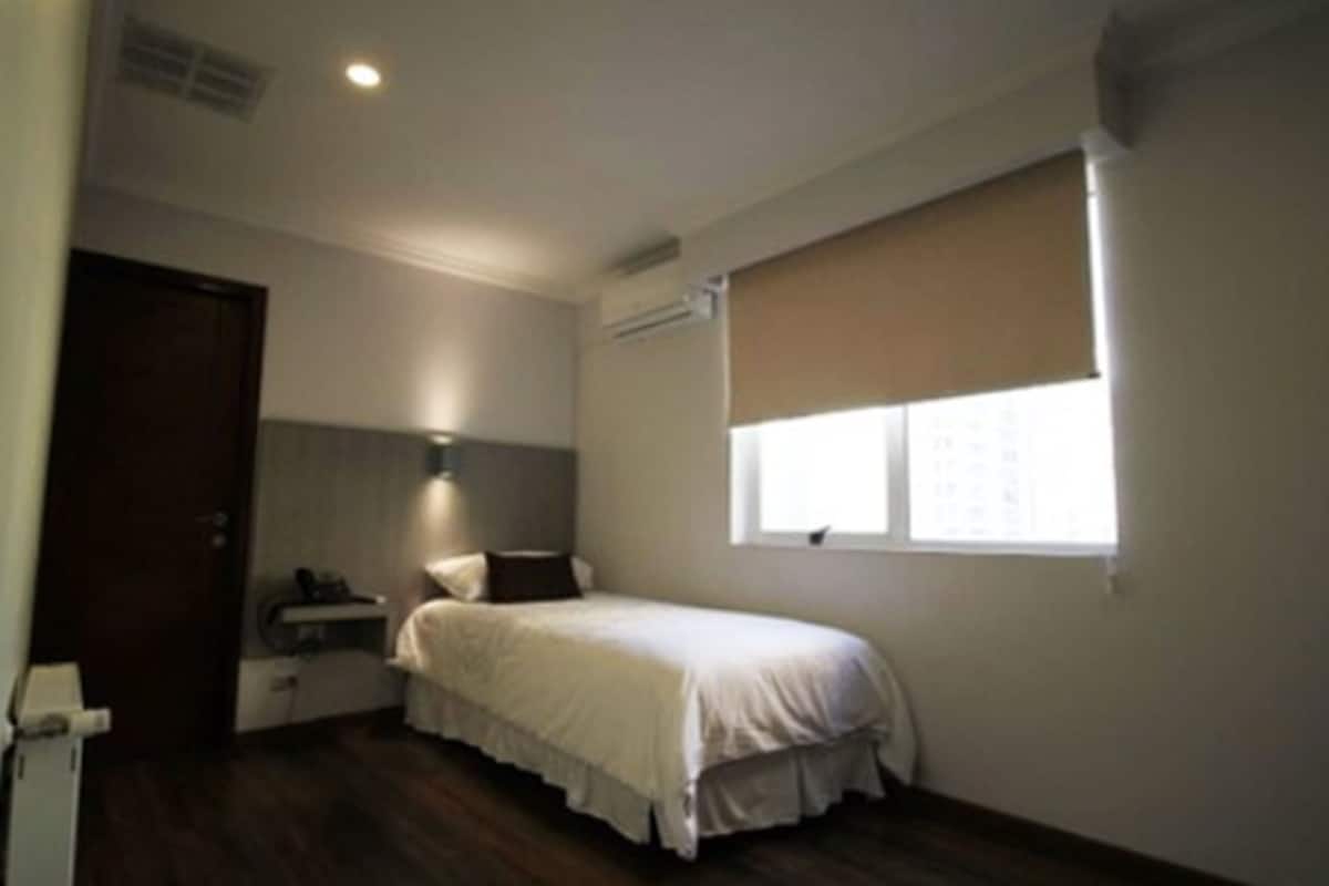 Ein kleines Zimmer mit einem Einzelbett, Holzboden, einem Nachttisch, einem Fenster mit Jalousien und einer Tür.