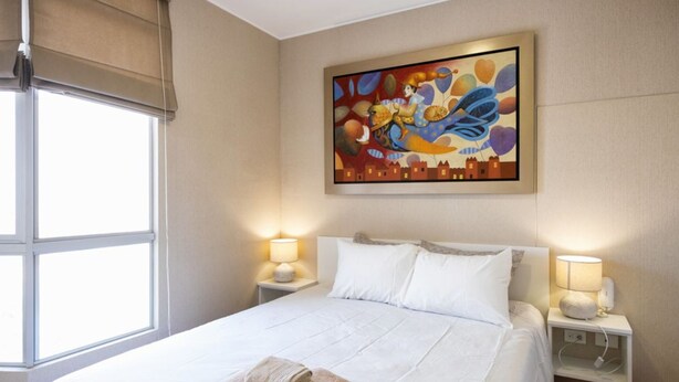 A hotel room with a large bed, two bedside tables with lamps, a window with blinds, and a colorful painting on the wall. - UR Place Rentals - 3Bdrm Av Brasil Lima (Lima)