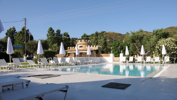 A pool area with white lounge chairs and umbrellas, a stone building in the background, and a clear blue sky. - Vicky's Garden (Zakynthos)