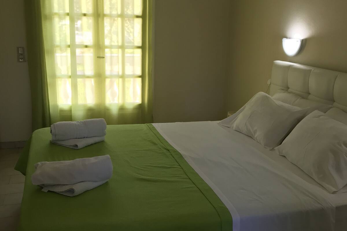 A neatly made bed with a green bedspread and white linens, a window with sheer curtains, and a wall-mounted light fixture.