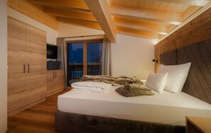 A bedroom with a wooden ceiling, a bed with white linens, a television on a stand, and a sliding glass door leading to a balcony. - NATURPARKGUT - alpdeluxe Apartments (Holzgau)