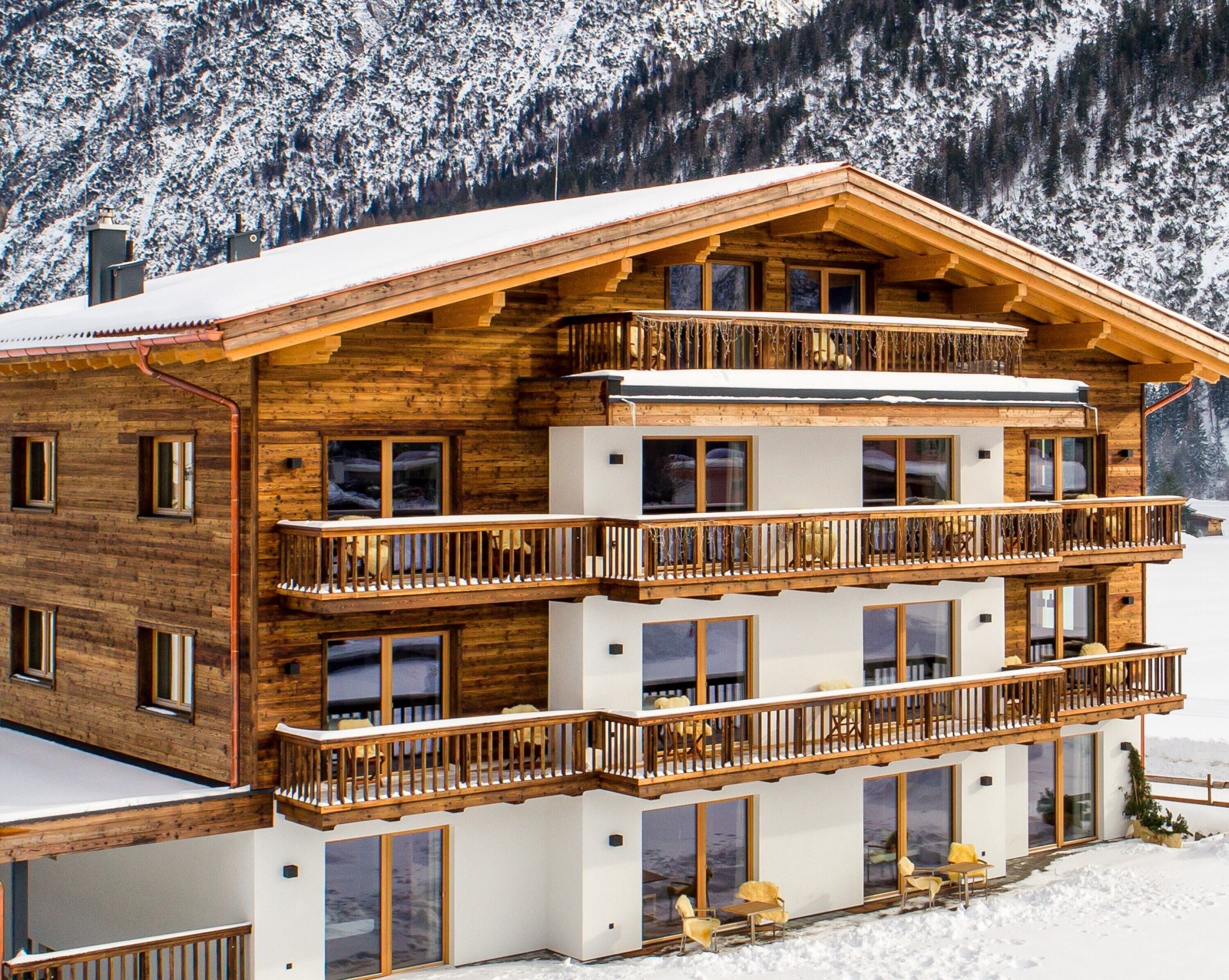 A multi-story wooden chalet with balconies, large windows, and a snowy mountain backdrop.