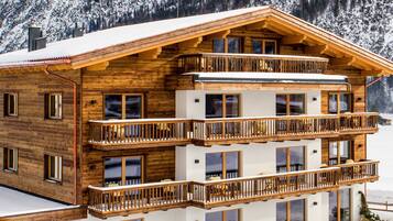 A multi-story wooden chalet with balconies, large windows, and a snowy mountain backdrop.