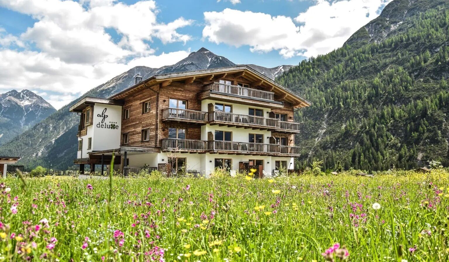 A wooden chalet with balconies, set in a mountainous area with a meadow in the foreground.
