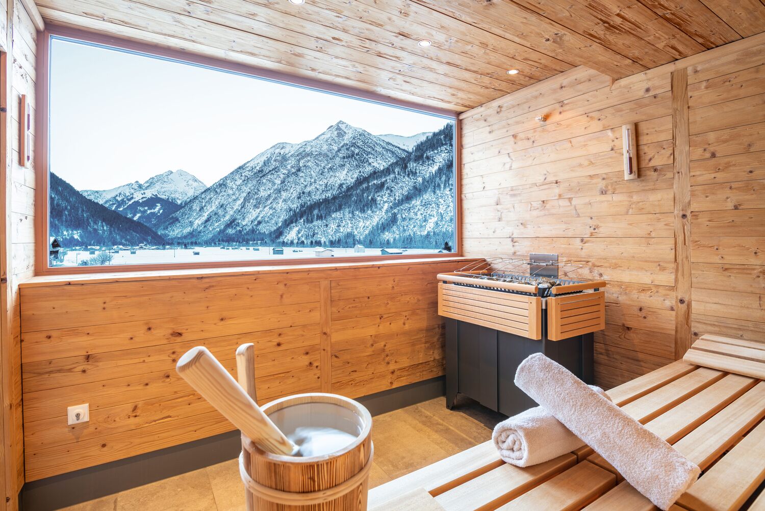 A wooden cabin with a large window overlooking snow-covered mountains. Inside, there is a wooden tub, a wooden bench, and a towel.