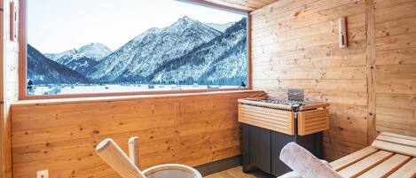 A wooden cabin with a large window overlooking snow-covered mountains. Inside, there is a wooden tub, a wooden bench, and a towel.
