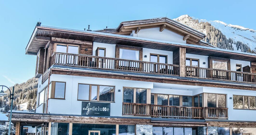 A multi-story hotel with balconies and large windows, set against a mountainous backdrop.