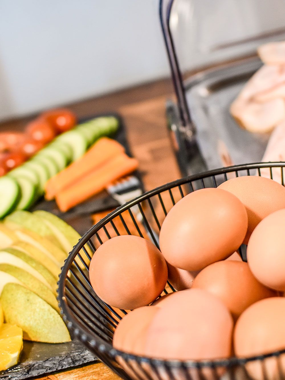 A kitchen counter with a basket of eggs, sliced cucumbers, carrots, and apples.