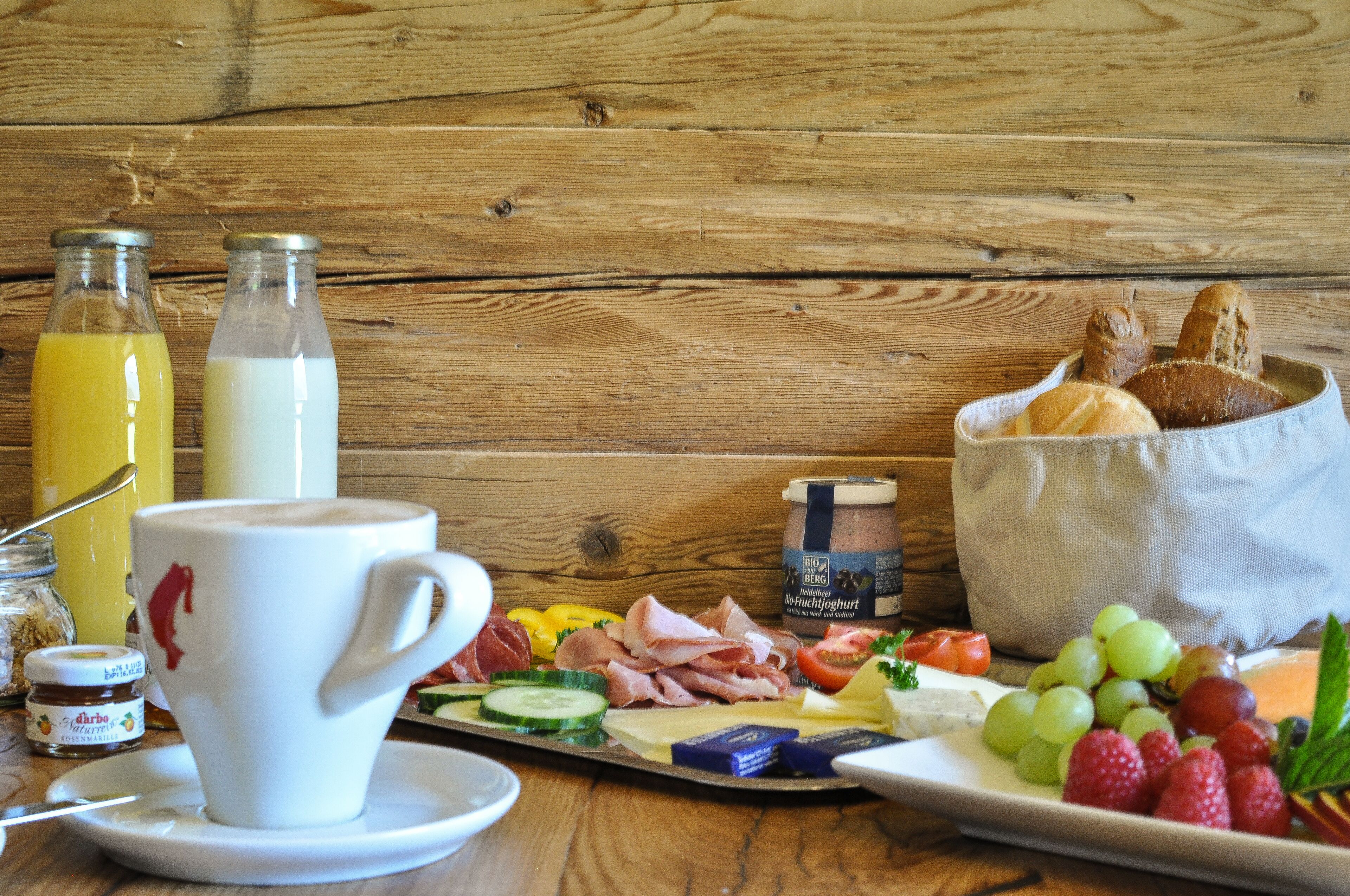 A breakfast table with a cup, bottles, bread, cheese, meats, fruits, and a jar of jam.