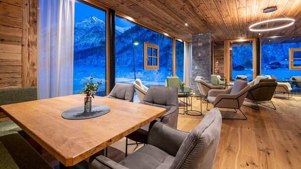 A cozy room with a wooden ceiling, large windows with a snowy mountain view, a wooden table with chairs, and a stone pillar.