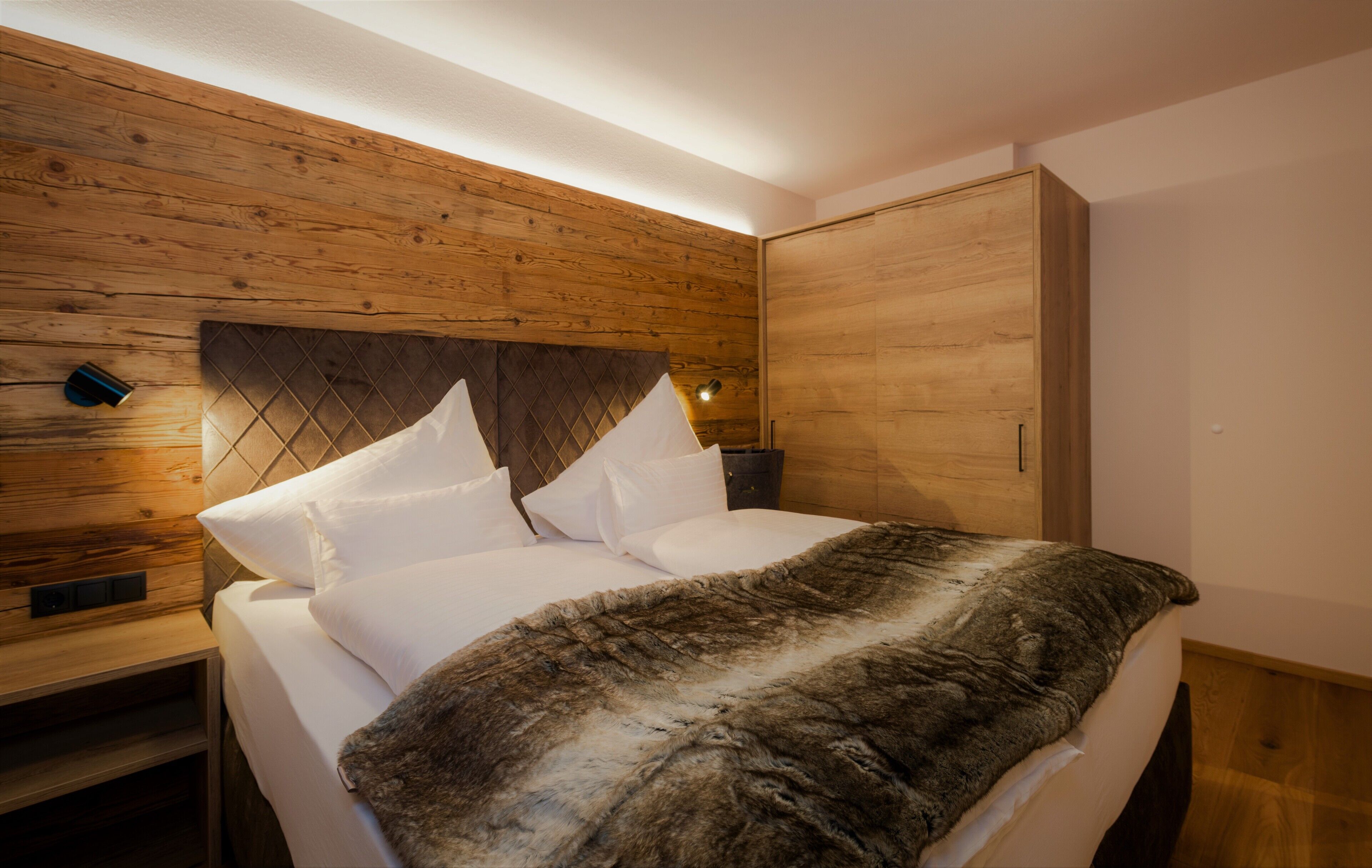Un dormitorio con cabecera de madera, cama con ropa blanca, un plaid de piel y una mesita de noche con lámpara.