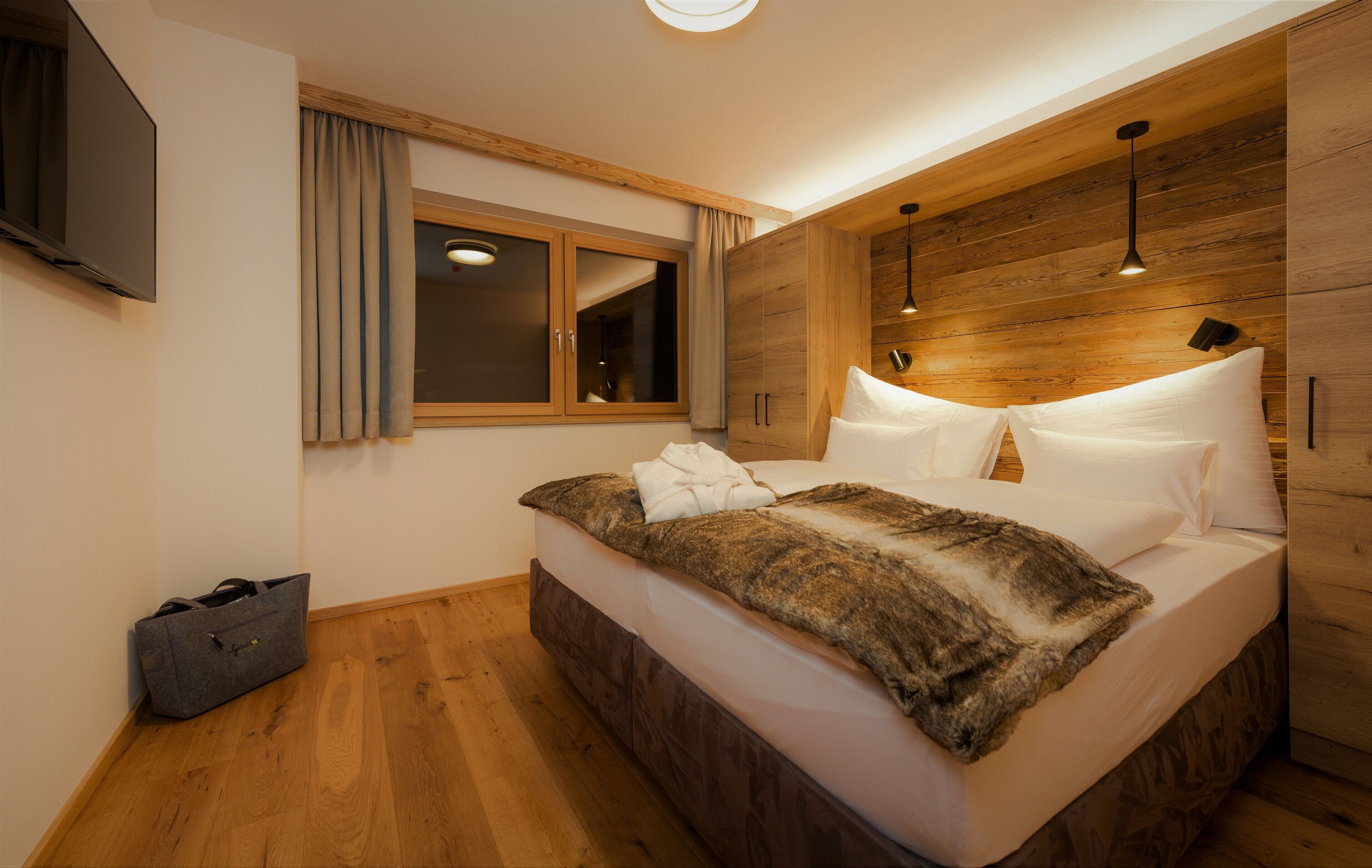 Une chambre à coucher moderne avec une tête de lit en bois, un grand lit avec du linge de lit blanc, une fenêtre avec des rideaux et un téléviseur à écran plat fixé au mur.