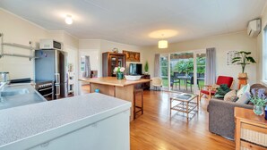 A modern kitchen with a built-in microwave, refrigerator, and sink. There is a wooden dining table with chairs, a sofa, and a coffee table in a living area with a view of the outdoors. - Sunny Grey Lynn Holiday Home (Auckland)