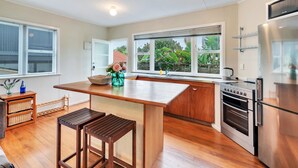 A modern kitchen with wooden flooring, a central island, stainless steel appliances, and large windows offering a view of greenery. - Sunny Grey Lynn Holiday Home (Auckland)