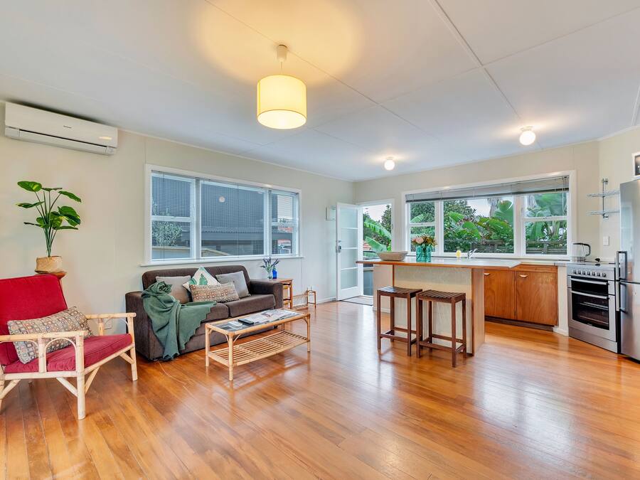A spacious living area with a wooden floor, a red chair, a sofa, and a coffee table. There is a kitchen with a refrigerator, oven, and a sink.