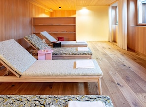 A sauna room with wooden floors, three sun loungers with towels, a small table with a pink top, and a shelf with towels.