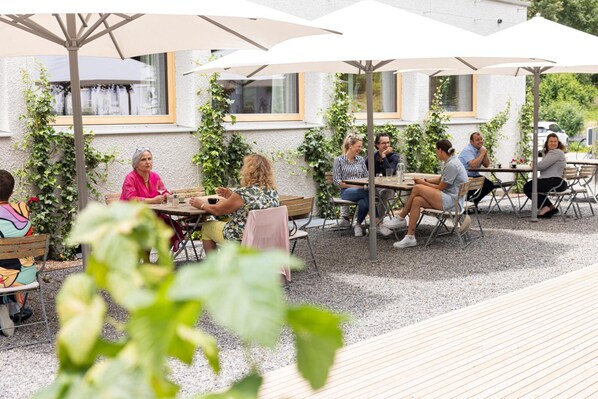 An outdoor seating area with tables and umbrellas, surrounded by greenery and a building with windows.