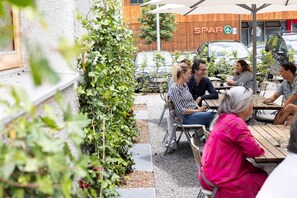 An outdoor seating area with tables and chairs, surrounded by greenery and a building with a wooden facade.