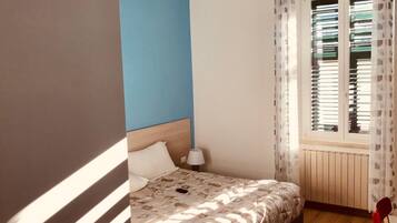 A bedroom with a bed, a nightstand, a window with blinds, and a view of the outside.