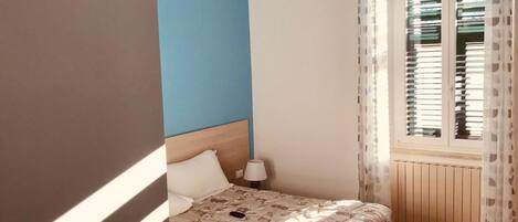 Un dormitorio con cama, mesita de noche, ventana con persianas y vista al exterior.