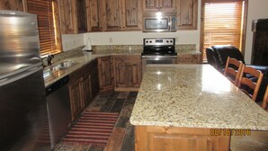 A kitchen with wooden cabinets, a granite countertop, stainless steel appliances, and a dining area with chairs. - Townhomes on the Green (Afton)