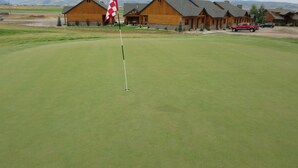 A golf course with a flagstick in the foreground and wooden cabins in the background. - Townhomes on the Green (Afton)