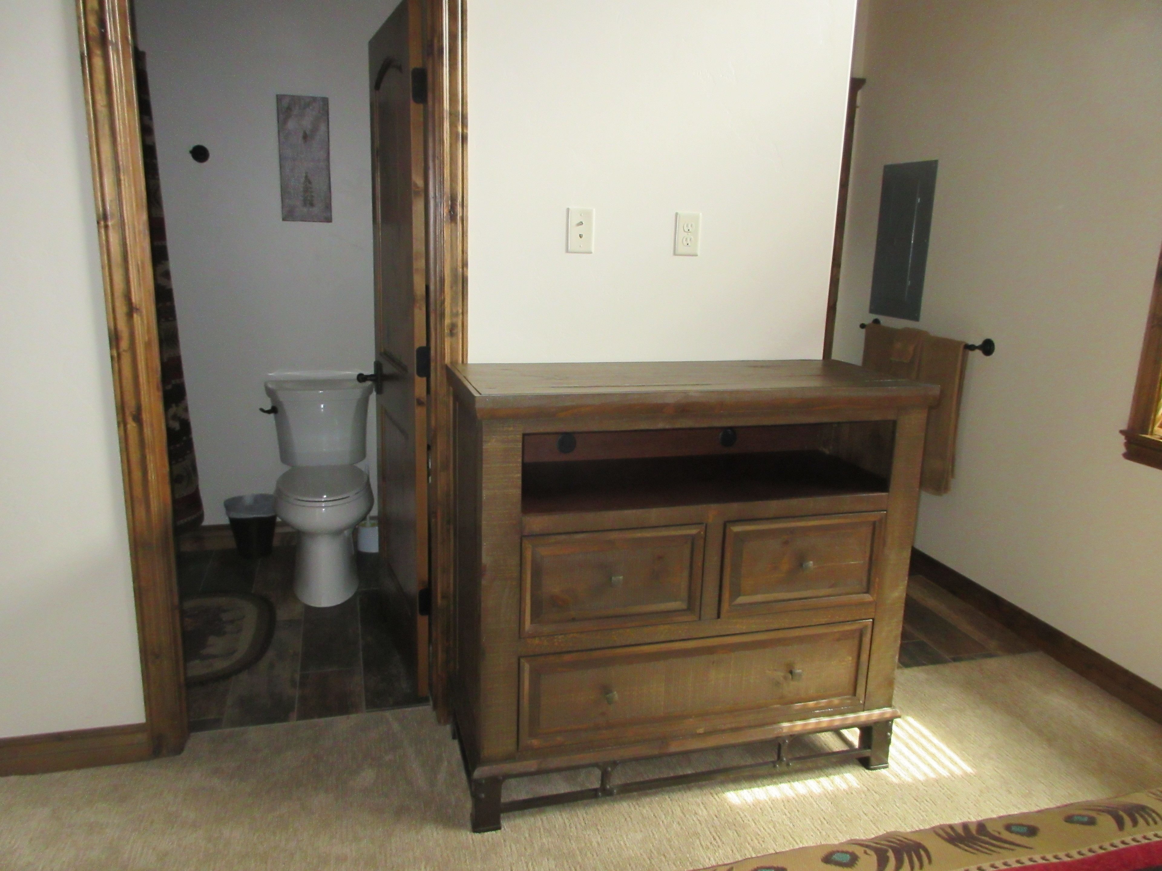 A wooden dresser with drawers, a mirror, and a small shelf in a room with a toilet and a rug.