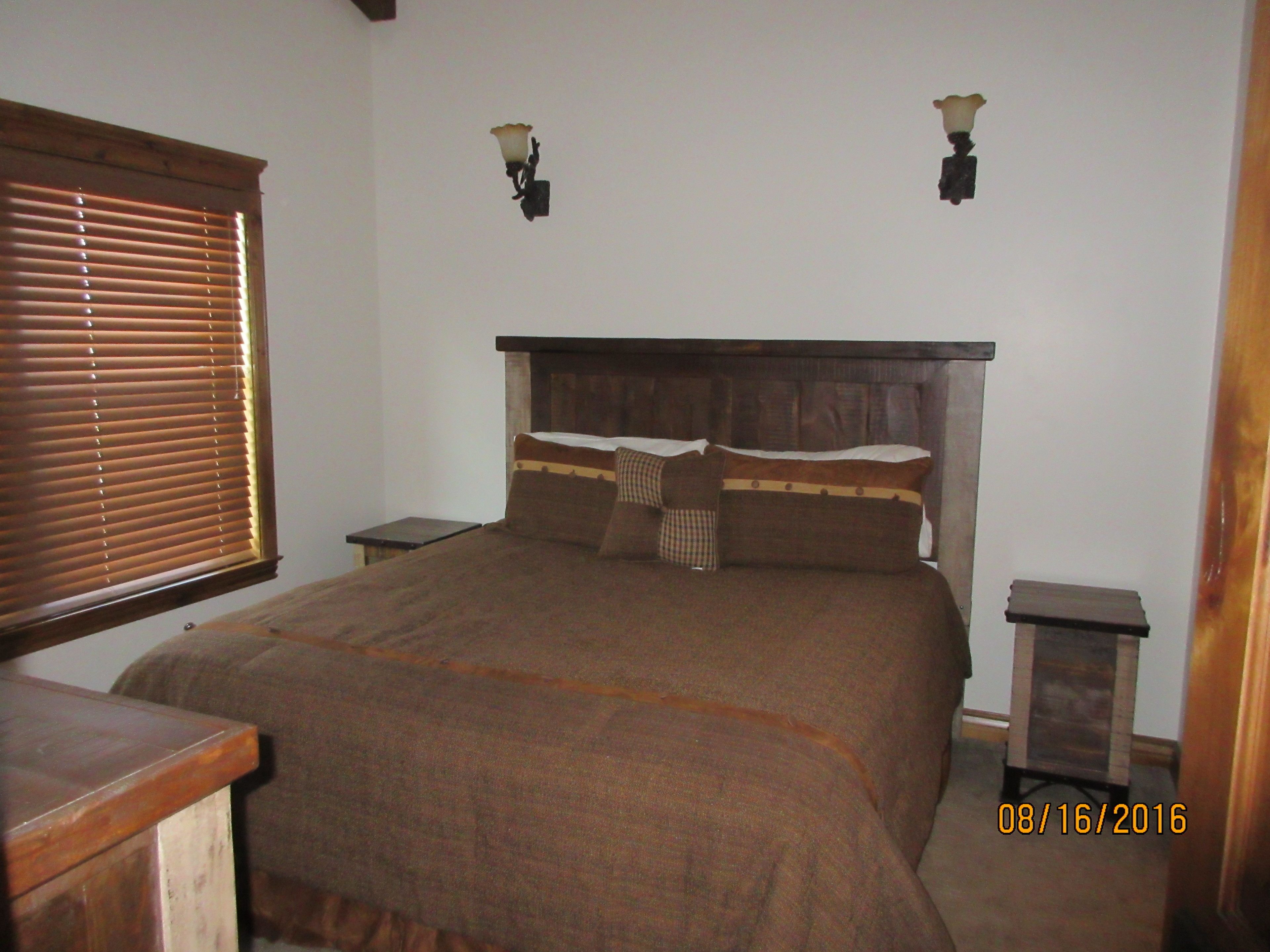 Un dormitorio con cama de madera, dos mesitas de noche, una ventana con persianas y lámparas de pared.