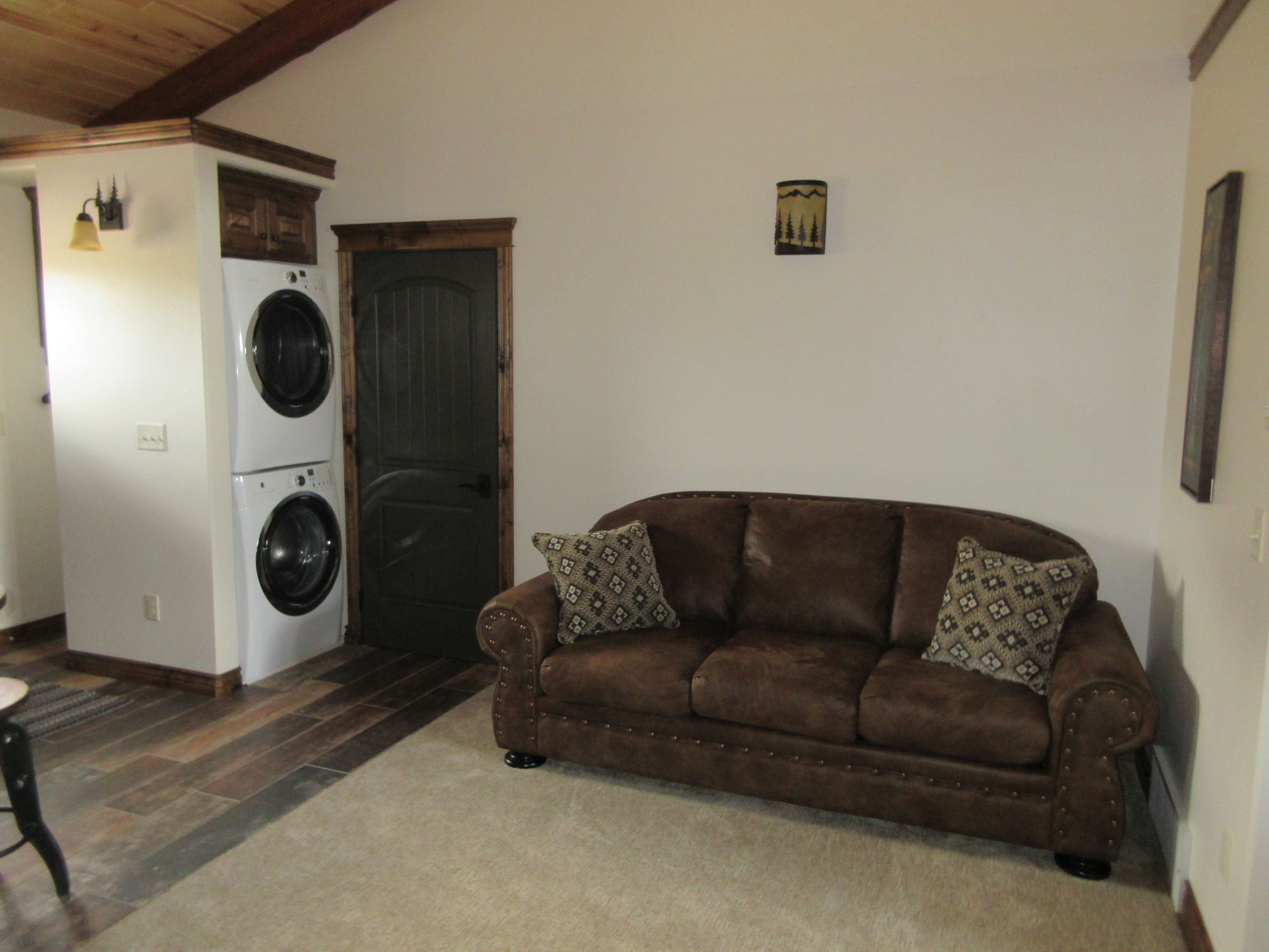 Een woonkamer met een bruine bank, gekleurde kussens, een wasmachine en een deur.
