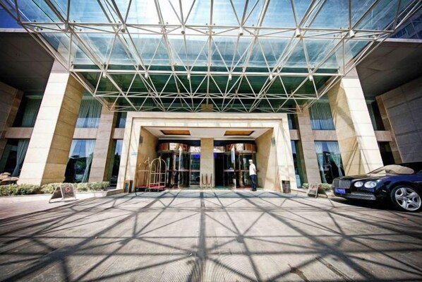 A hotel entrance with a glass canopy, a car parked outside, and a person walking towards the entrance. - Sizhou Hotel (Xuyi)
