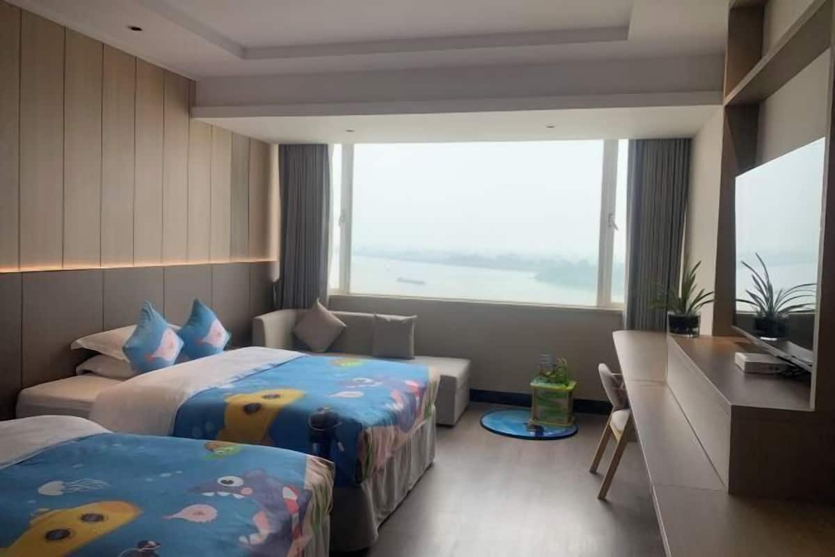 A hotel room with two beds, a desk, a chair, a TV, and a large window with a view.