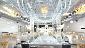 Un'ampia sala con un palco centrale, file di tavoli bianchi con decorazioni dorate e un lampadario.