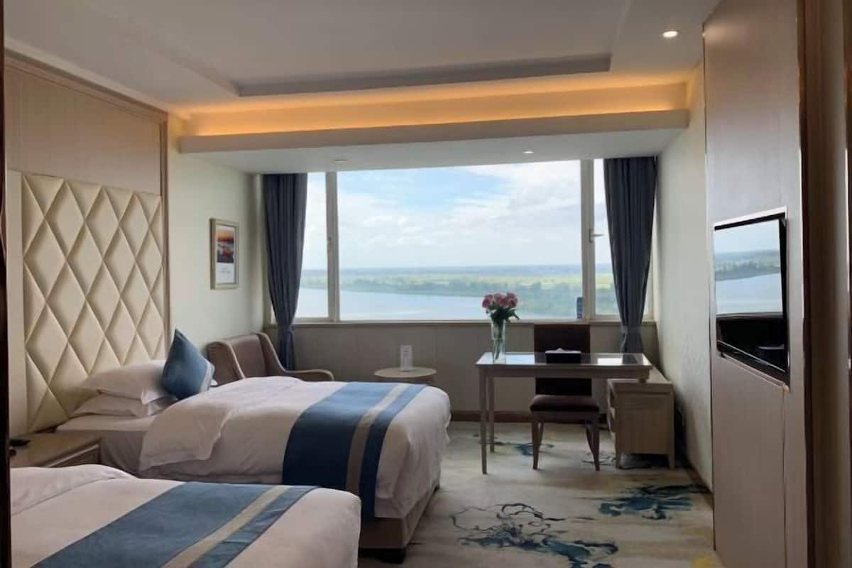 A hotel room with two beds, a desk, a TV, and a large window with a view of a lake.