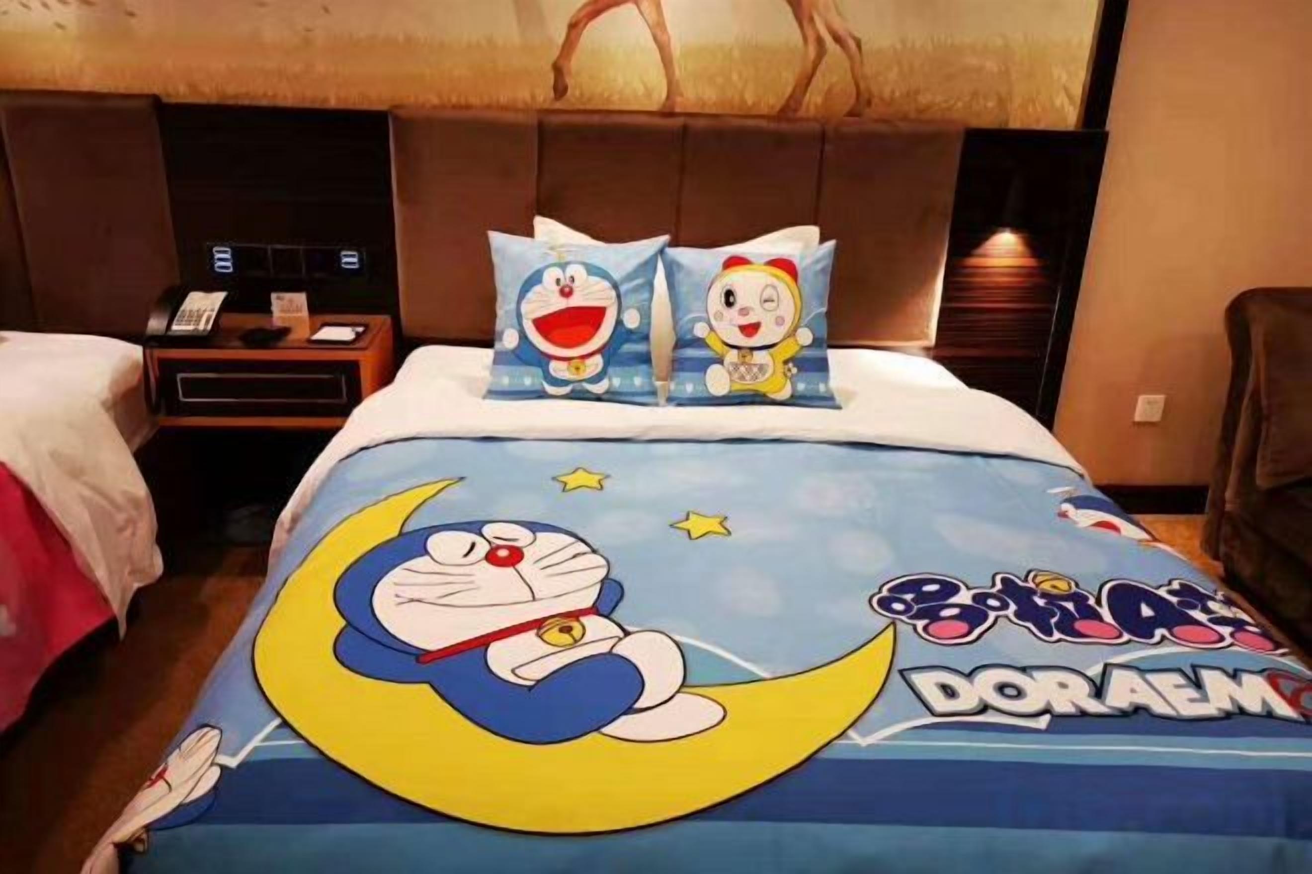 A hotel room with a bed featuring a Doraemon-themed bedspread, a nightstand with a telephone, and a headboard with a light fixture.