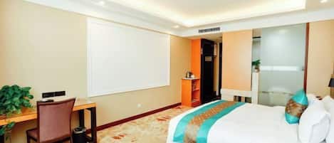 A hotel room with a bed, a desk, a chair, a small table, a plant, and a mirror.