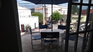 An outdoor patio with a table and chairs, a blue umbrella, and a view of the street and buildings in the background.