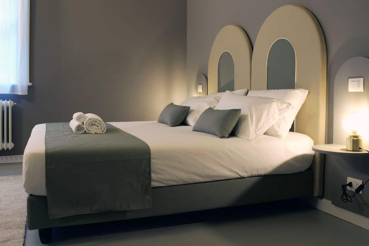 A neatly made bed with a green blanket and rolled towels, a bedside table with a lamp, and a radiator in the background.