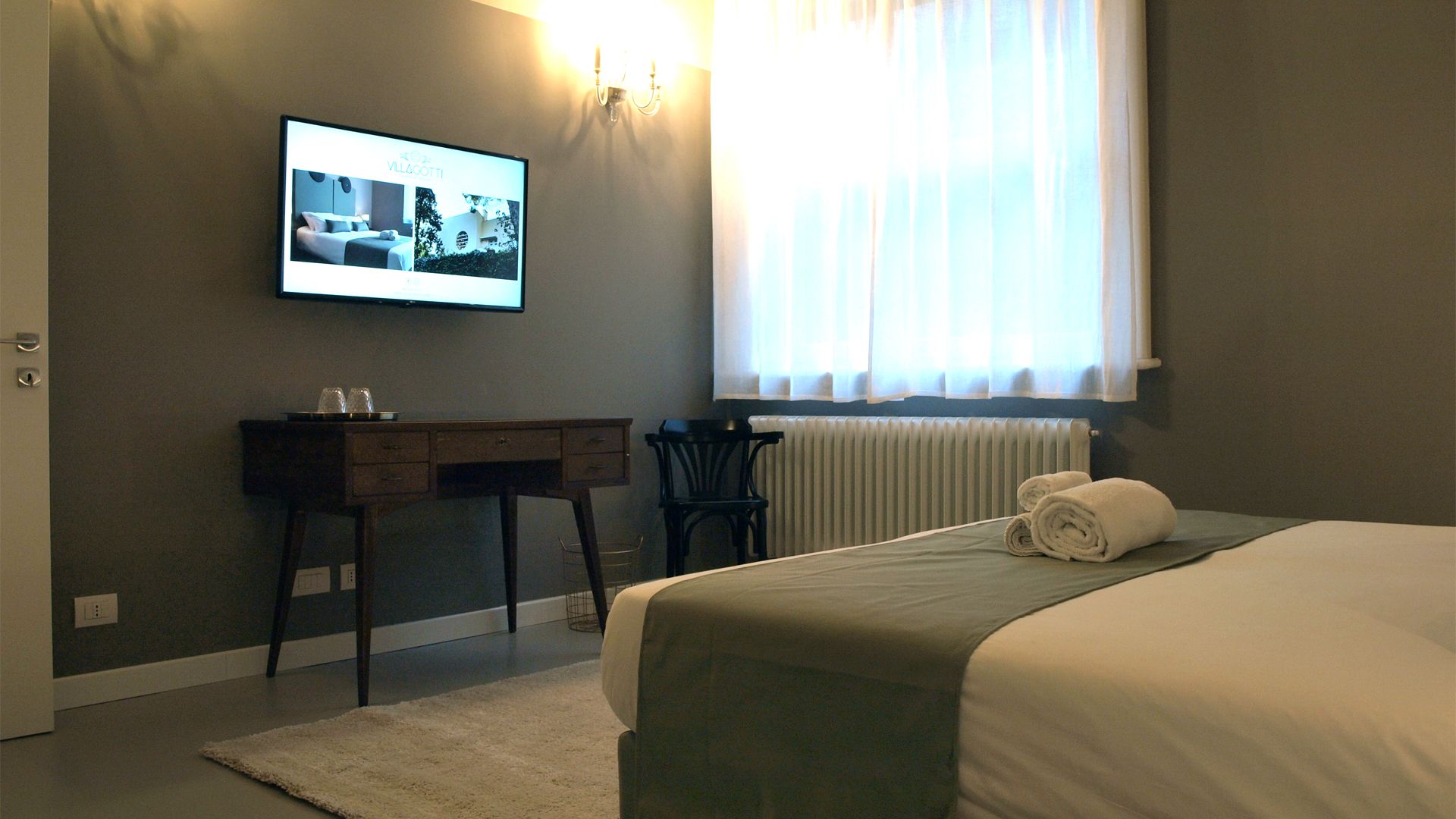Habitación de hotel con cama, escritorio, silla, radiador y ventana con cortinas.
