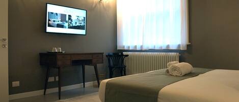 Habitación de hotel con cama, escritorio, silla, radiador y ventana con cortinas.