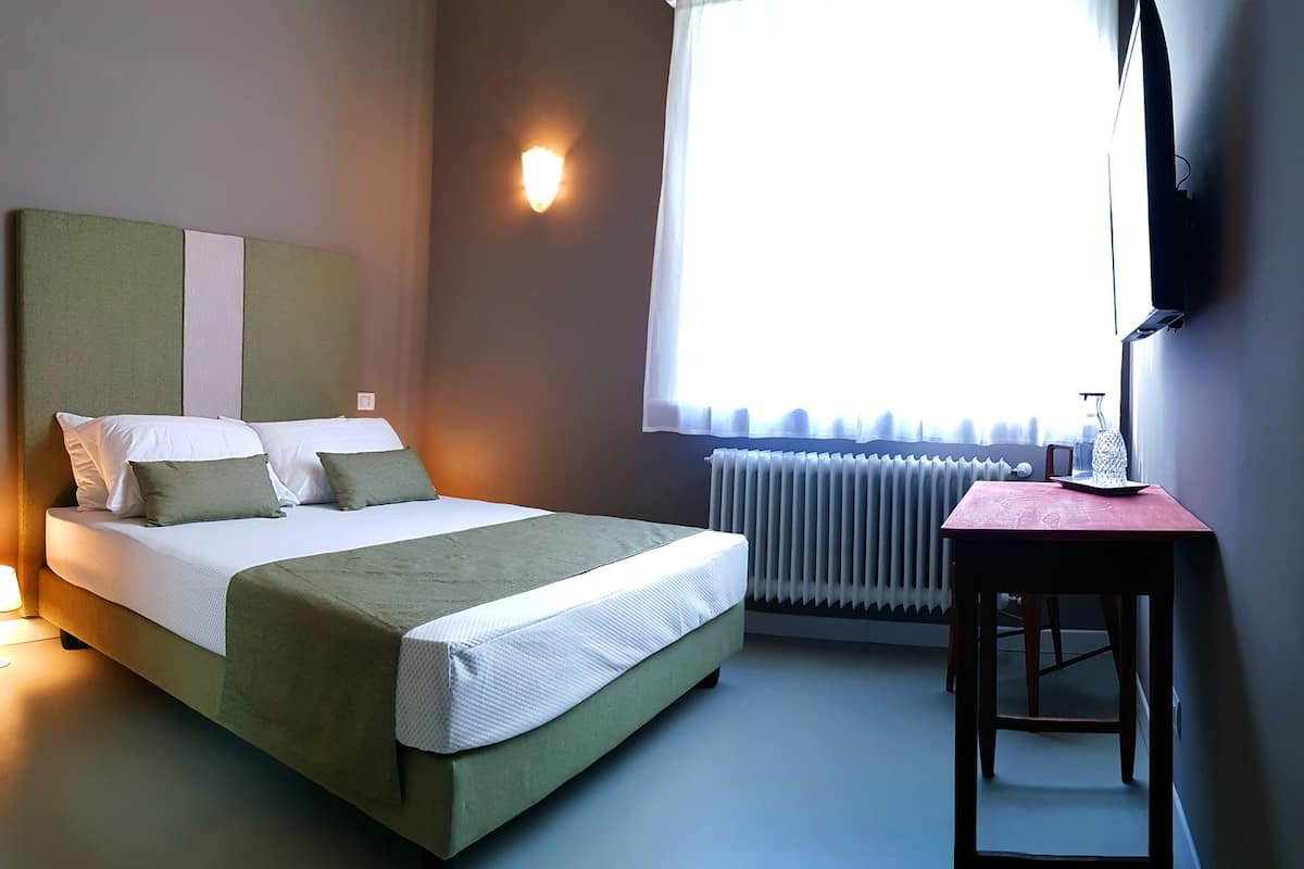 A neatly made bed with pillows and a blanket, a bedside table with a lamp, a radiator, and a window with curtains.