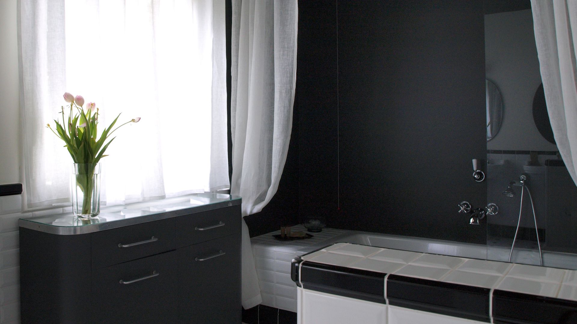 A modern bathroom with a dark tiled wall, a white bathtub, and a glass vase with flowers on a vanity.