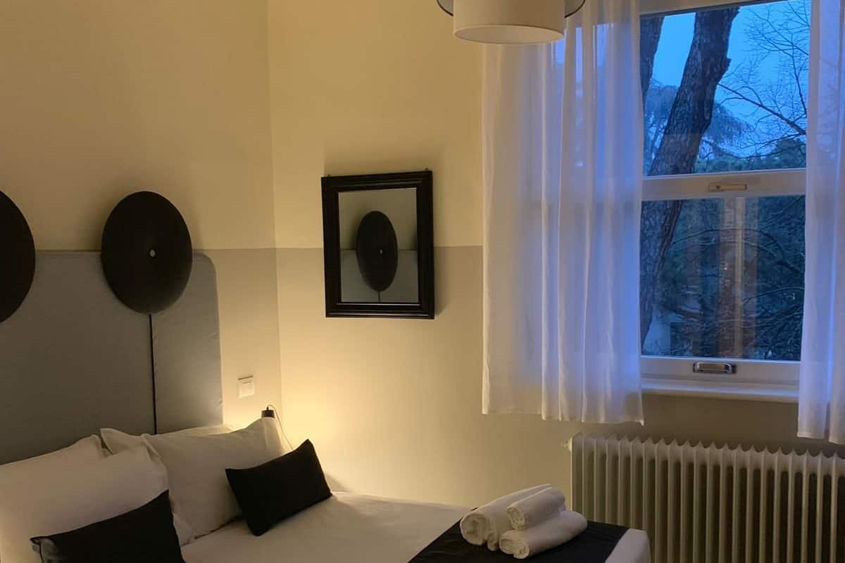 A modern hotel room with a large bed, a bedside table, a hanging light fixture, a window with curtains, and a radiator.