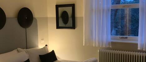 Una habitación de hotel moderna con una cama grande, una mesita de noche, una lámpara colgante, una ventana con cortinas y un radiador.