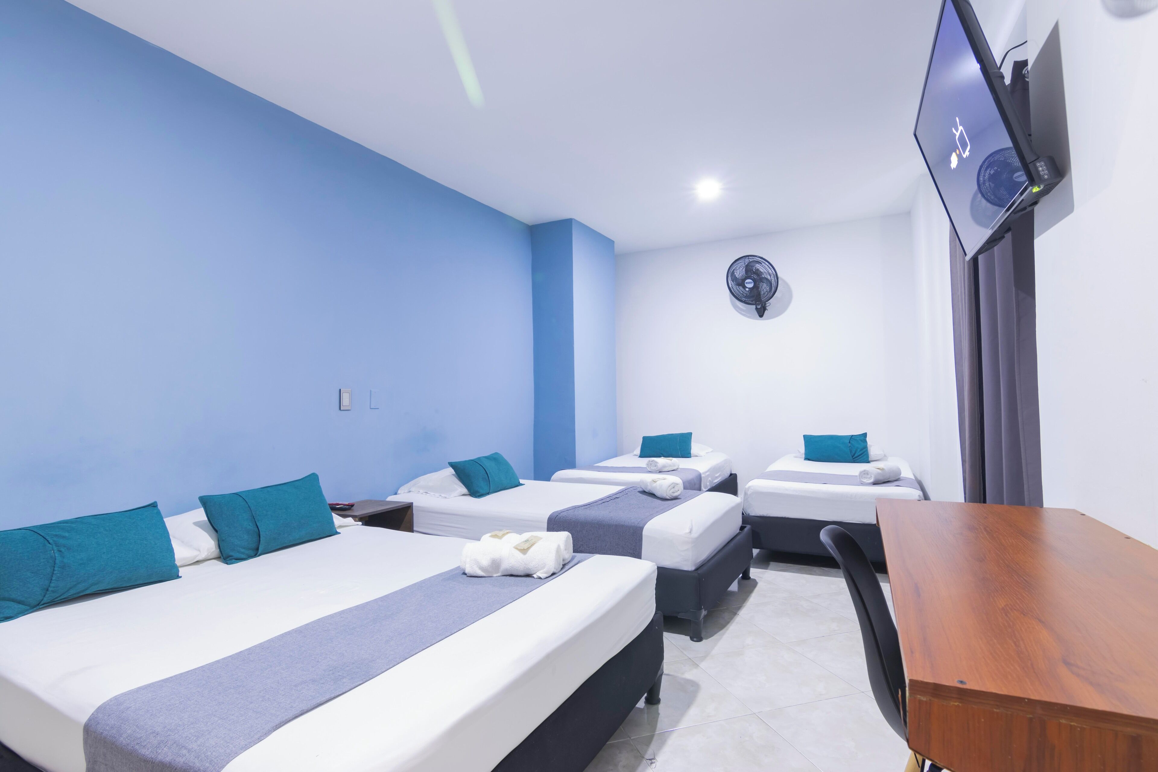 Quarto de hotel com três camas, uma escrivaninha, televisão e ventilador.