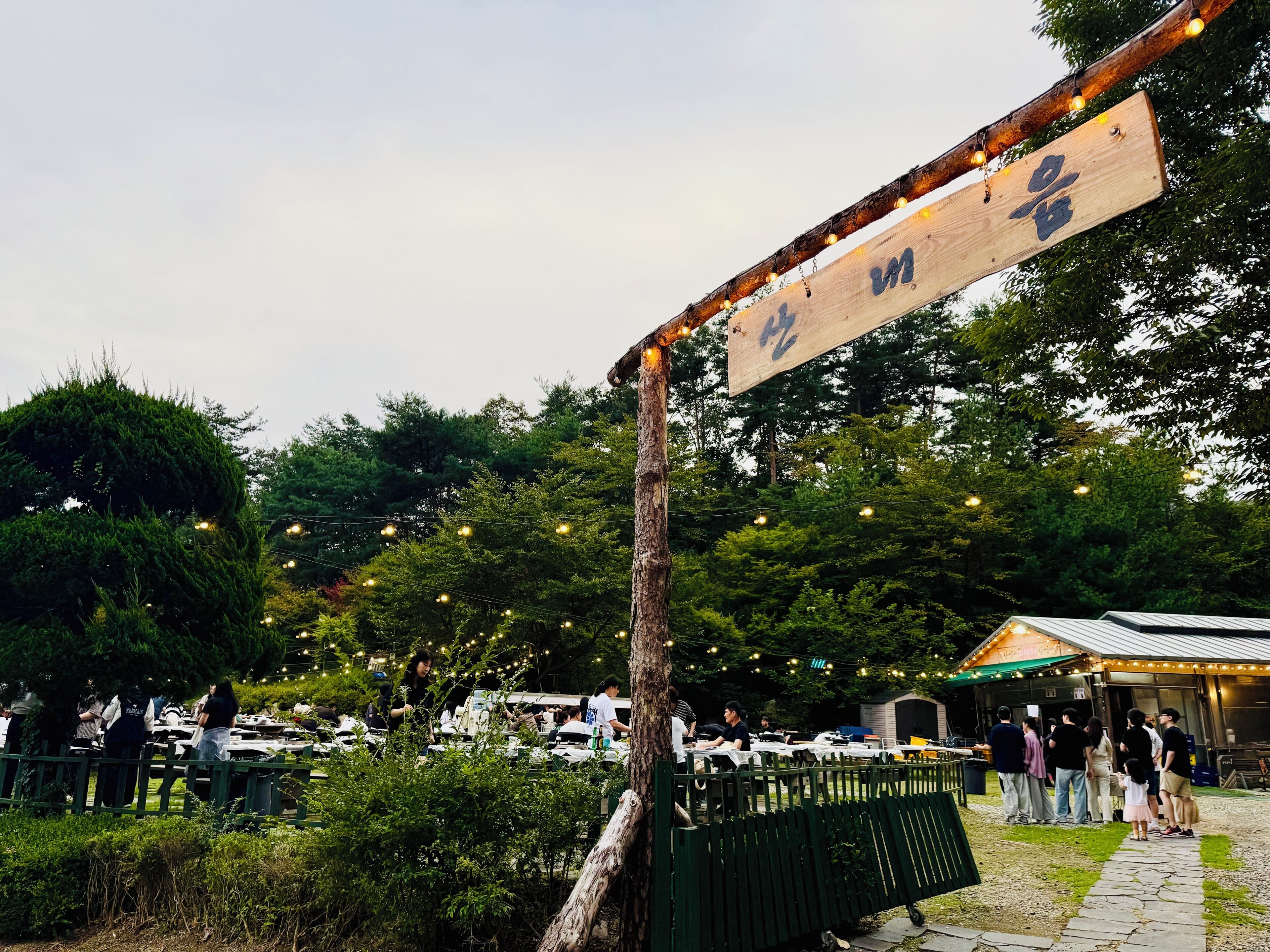 An outdoor dining area with tables and chairs, surrounded by greenery and a wooden sign with characters.
