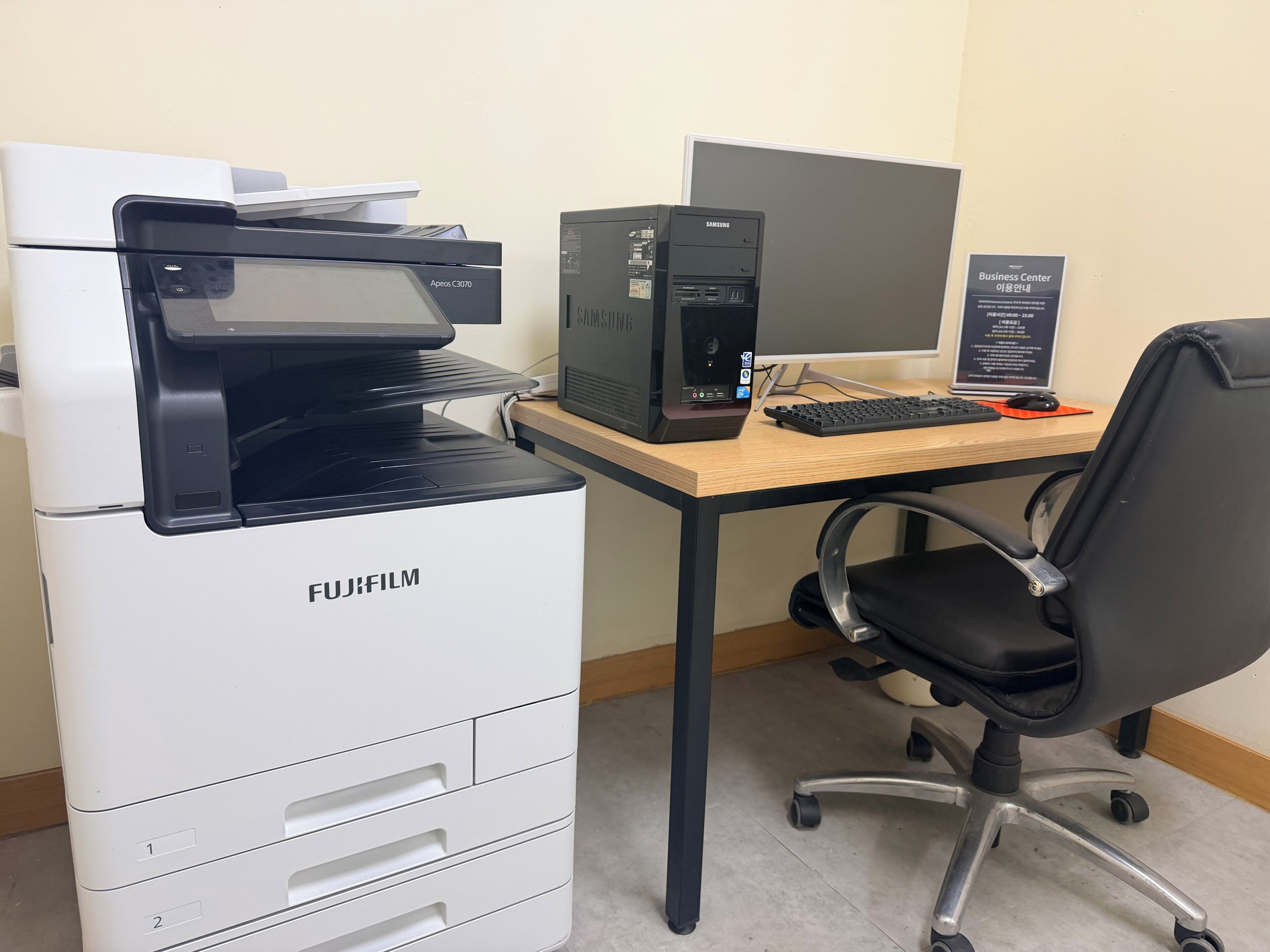 Ein FUJIFILM-Drucker, ein Samsung-Desktop-Computer, eine Tastatur, eine Maus und ein schwarzer Bürostuhl.