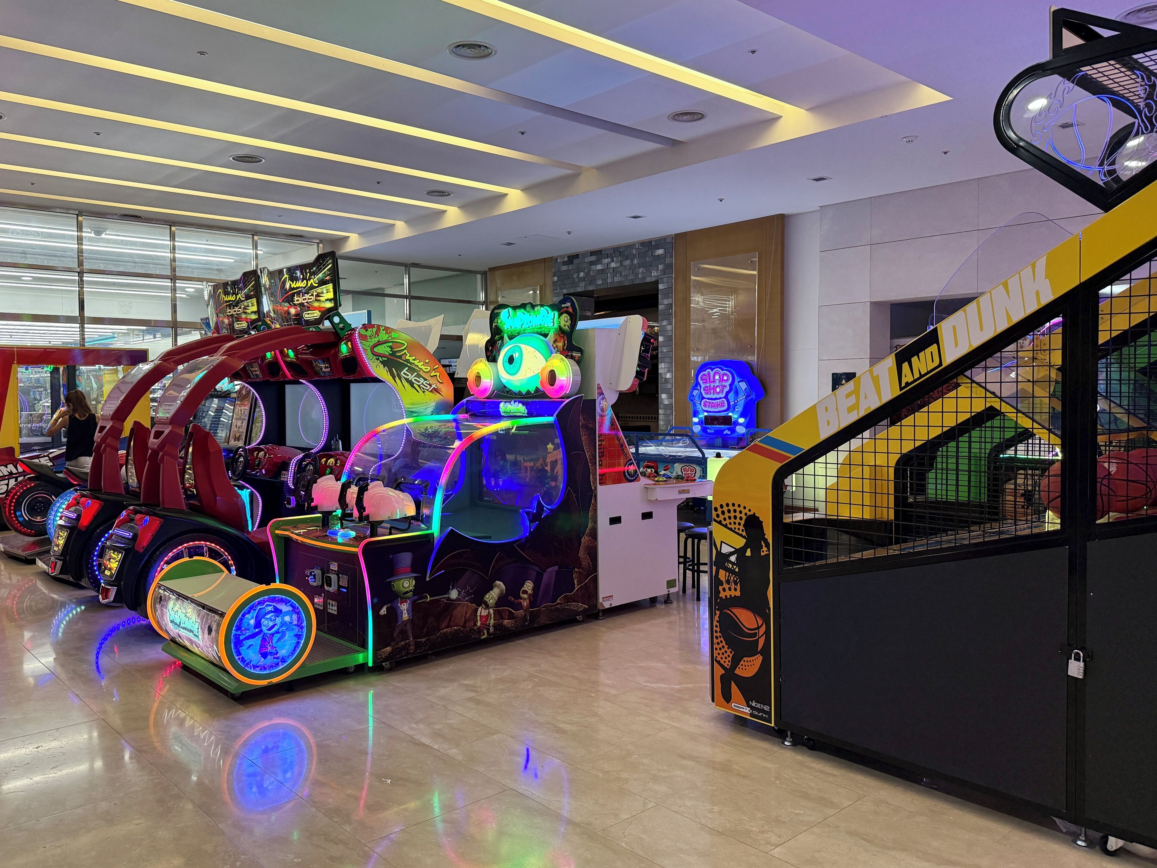 Um salão de jogos indoor com diversas máquinas de entretenimento, uma escadaria visível e um letreiro de neon.