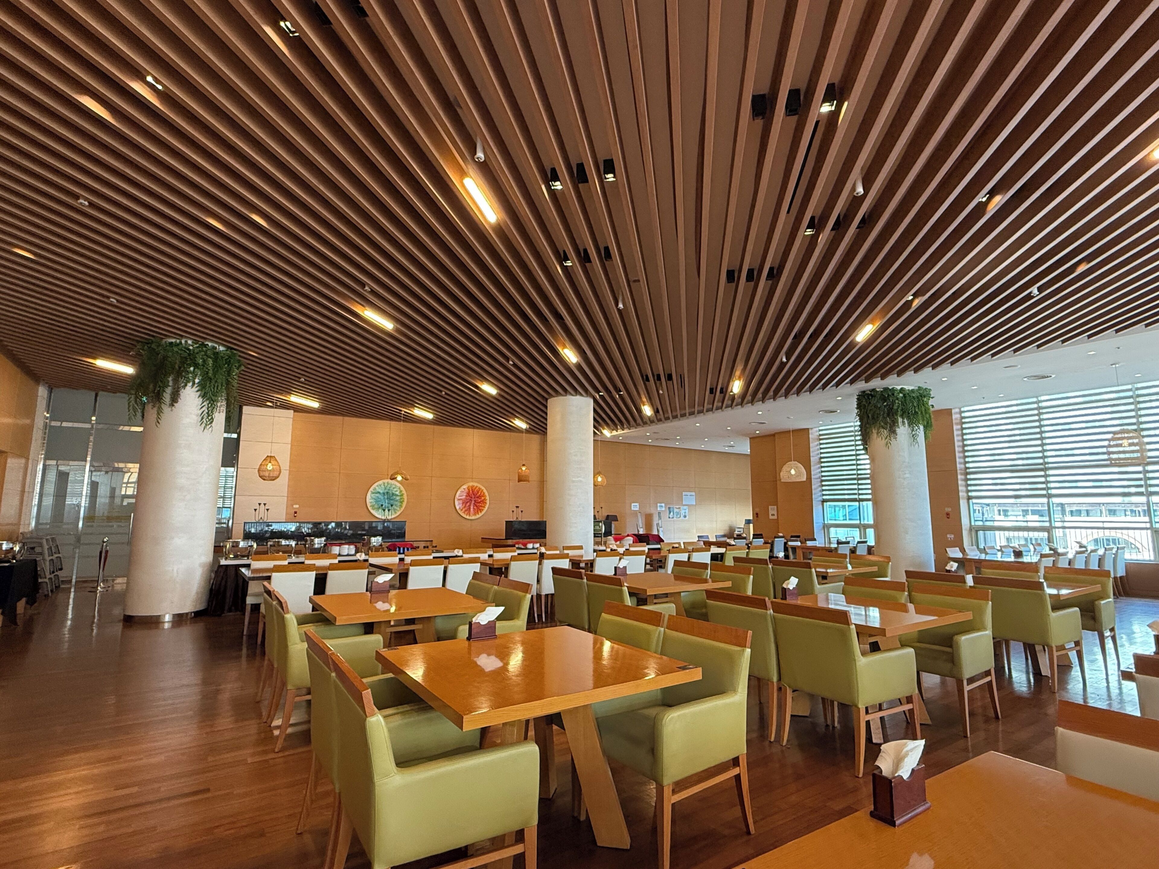 A spacious restaurant with wooden ceiling panels, green cushioned chairs, and wooden tables.
