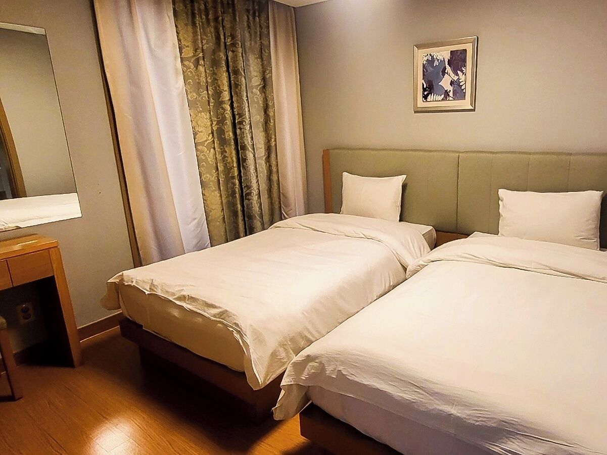 Quarto de hotel com duas camas, uma mesinha de cabeceira de madeira, um espelho e um quadro na parede.